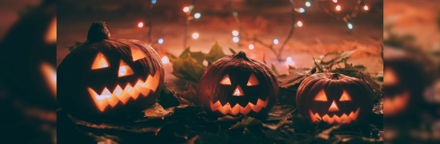 Three illuminated carved pumpkins on a table covered in fairy lights and autumnal leaves.