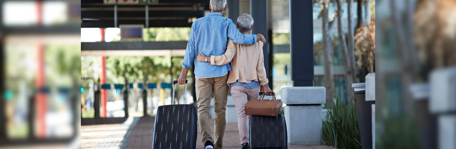 A grey-haired couple with one arm around each other carrying suitcases and walking towards the airport exit.